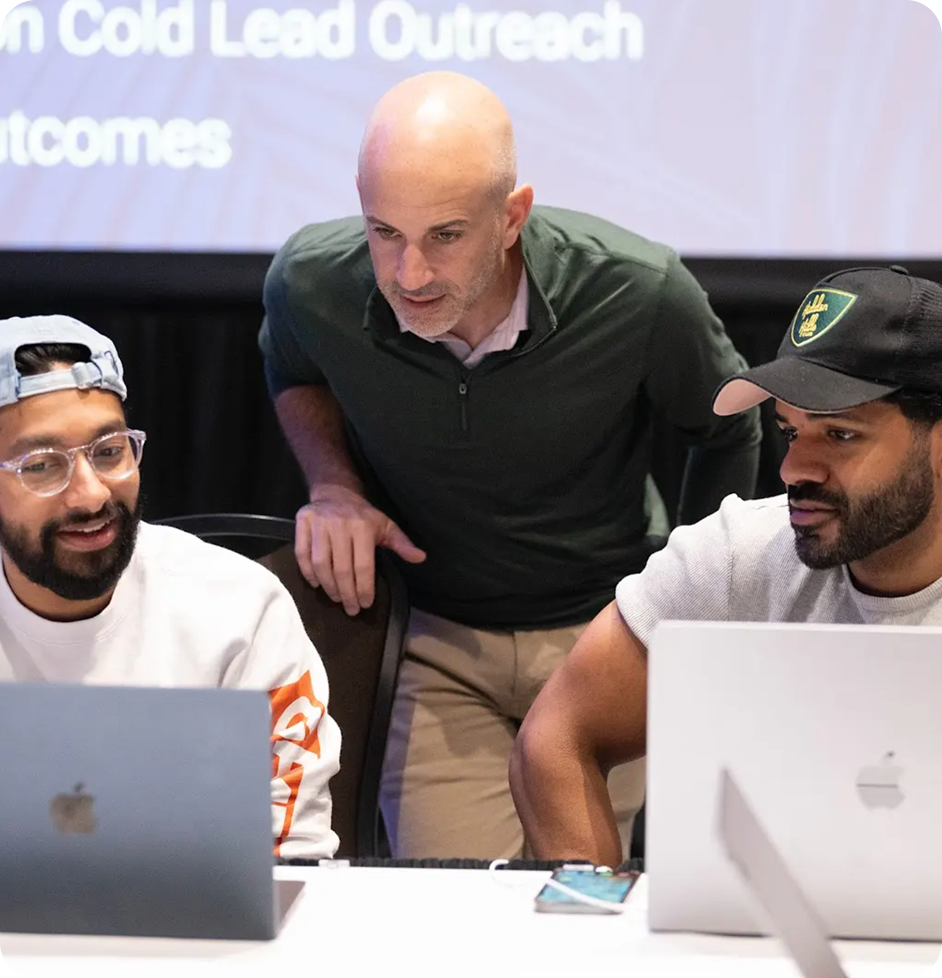 Two men sitting down at a desk with laptops while a third man is standing behind them.